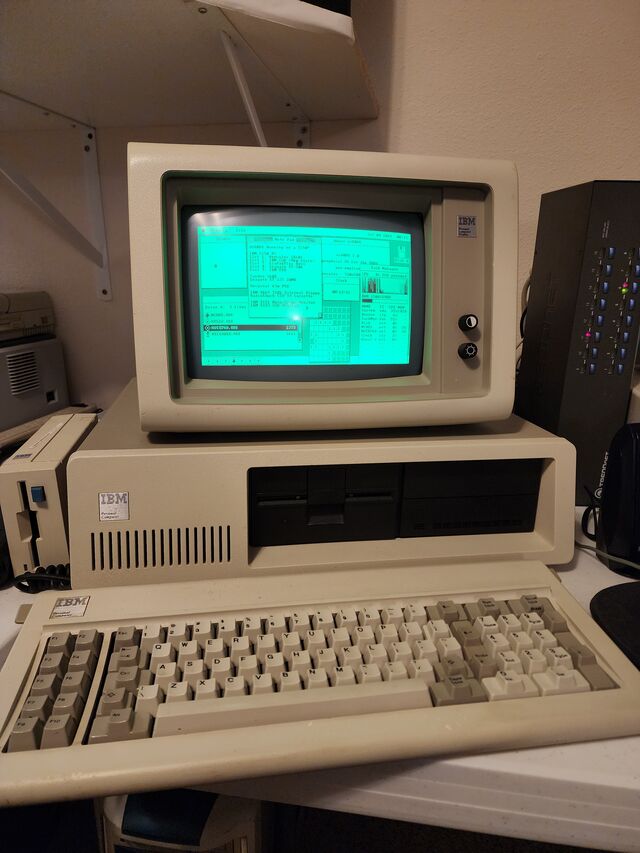 An IBM 5150 Personal Computer with its Model F keyboard in front and an IBM 5151 monochrome monitor sitting on top of the case. The green phosphor screen shows the os8088 desktop: a menu bar, several overlapping windows and a dock along the bottom.