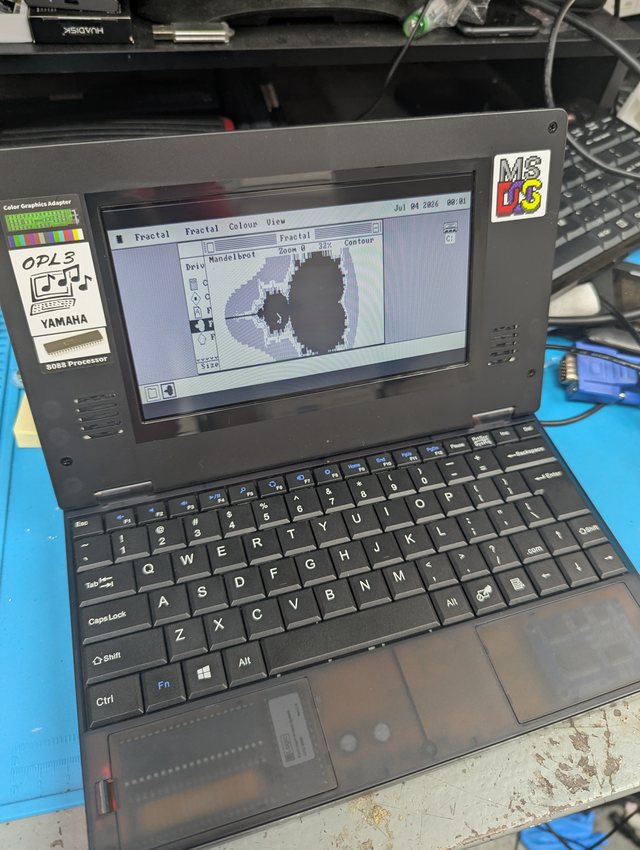 A small black Book8088 laptop open on a workbench, with stickers on the lid reading Color Graphics Adapter, OPL3 Yamaha, 8088 Processor and MS-DOS. Its wide LCD shows the os8088 desktop in CGA: a menu bar reading Fractal, Fractal, Colour and View with the date Jul 04 2026 on the right, a Fractal window drawing a black and white Mandelbrot set at Zoom 0 and 32 percent, a Disk window behind it, and a C drive icon in the top right corner.